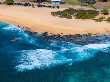 Hawaii, Oahu adasındaki bir kumsalın havadan görünüşü, altın kum, turkuaz sular, kayalık çıkıntılar, küçük bir bina ve park edilmiş arabalar..