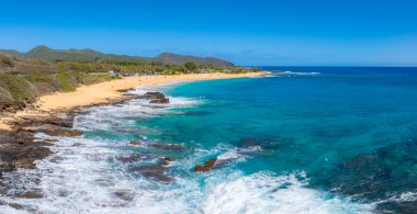 Hawaii, Oahu adasındaki bir kumsalın havadan görünüşü, altın kum, turkuaz sular, kayalık çıkıntılar, yeşil tepeler ve park halindeki araçların bulunduğu küçük bir yol..