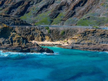 Bereketli yeşilliği olan engebeli uçurumlar Oahu adasında canlı turkuaz sularla karşılaşıyor. Uçurum kenarındaki yolun altında görünen araçlarla kumlu bir sahil var..