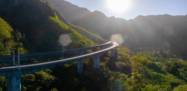 Oahu Adası, Hawaii 'deki yeşil dağların arasından geçen kıvrımlı, yüksek bir otoyol. Güneş ışığı, dramatik bir dağ zeminiyle bitki örtüsünü ön plana çıkarıyor..