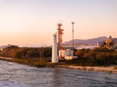 Hawaii, Oahu adasındaki sahil manzarası, beyaz bir deniz feneri, kırmızı kule, iletişim direği, kayalık kıyı şeridi, yeşillik ve dağ silueti..