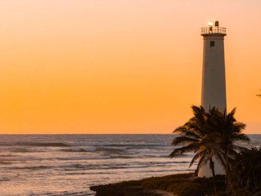Palmiye ağaçlarıyla çerçevelenmiş, kıyı uçurumunda uzun beyaz bir deniz feneri, gün batımında okyanusa bakıyor. Gökyüzü turuncu parlıyor, mavi suyla çelişiyor..