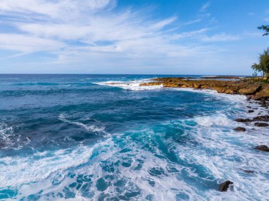 Canlı mavi okyanus dalgaları Oahu, Hawaii 'deki kayalık bir kıyı şeridine çarpıyor. Parçalı bulutlu bir gökyüzünün altında, kıyı boyunca ağaçlar ve bitki örtüsüyle birlikte..