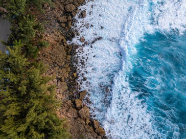 Oahu 'nun kıyı şeridinin yeşil ağaçlarla çevrili kayalık kıyılara çarpan turkuaz dalgaları ve sol tarafta dar bir yol görüntüsü..