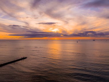 Oahu, Hawaii yakınlarında gün batımı, turuncu ve mor renkler, solda bir jet, insan siluetleri, yelkenliler, daha büyük bir gemi ve dramatik bulutlar..