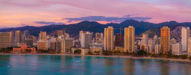 Modern binalar, Waikiki Sahili, turkuaz okyanus suları ve canlı günbatımı gökyüzü altında yemyeşil dağların yer aldığı Honolulu, Hawaii 'nin hava manzarası..