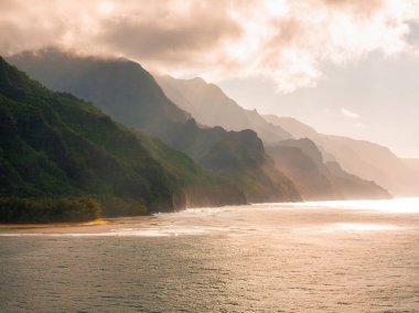 Hawaii, Kauai Adası 'ndaki Napali Sahili' nin sarp kayalıkları altın güneş ışığı, kumlu bir sahil ve sisli okyanus yansımaları ile kaplıdır..