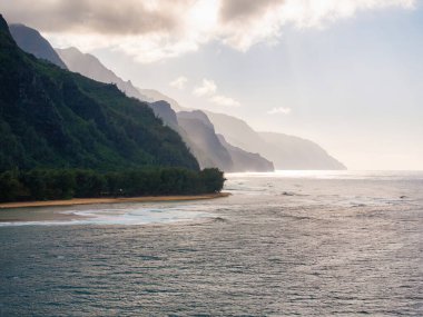 Napali sahilinin yemyeşil kayalıkları Pasifik Okyanusu 'na, sis, güneş ışığı, kumlu bir sahil ve ön planda yoğun bitki örtüsüyle iniyor..