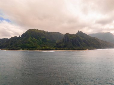 Havai 'deki Kauai Adası' nda bulutlu ve güneşli bir gökyüzünün karışımı altında, yumuşak dalgalarla dolu kumlu bir plajın üzerinde yemyeşil uçurumlar yükselir..