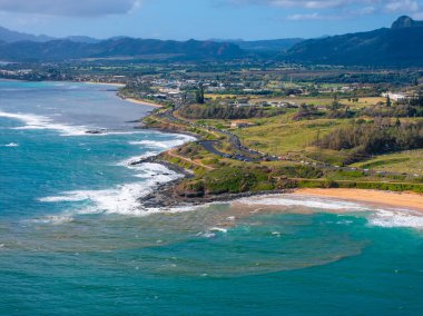 Hawaii, Kauai 'nin hava görüntüsü turkuaz suları, kumlu plajlar, dolambaçlı bir sahil yolu, yeşil alanlar ve bulutlu bir gökyüzünün altındaki uzak dağları gösteriyor..