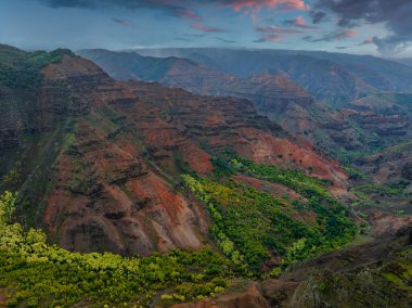 Hawaii, Kauai Adası 'ndaki Waimea Kanyonu' nda kırmızı ve kahverengi uçurumlar, yeşil bitki örtüsü, katmanlı kaya oluşumları ve dramatik pembe ve mavi bir gökyüzü bulunur..