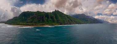 Hawaii, Kauai Adası 'ndaki Napali Sahili' nin hava görüntüsü engebeli yeşil uçurumları, kumlu bir sahili ve bulutlu bir gökyüzünün altında canlı mavi Pasifik Okyanusu sularını gösteriyor..
