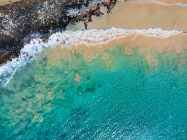 Altın kum, Kauai adasında turkuaz sularla birleşiyor. Dalgalar engebeli bir kayalık kıyıya çarpıyor ve uzaktaki mavi okyanus canlanıyor..