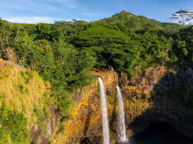 Wailua Falls cascades over a golden brown cliff in Kauai, Hawaii, surrounded by lush tropical vegetation, with a forested hill rising in the background. clipart