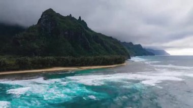 Bereketli yeşilliği olan dramatik Napali uçurumları kumlu bir sahile vuran okyanus dalgaları ve Hawaii, Kauai 'deki tepeleri kısmen kaplayan bulutlu bir gökyüzü..