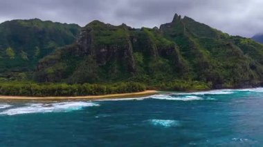 Hawaii, Kauai 'deki Napali kayalıklarının üzerindeki hava taraması, pürüzlü tepeler, yemyeşil, kumlu sahil, okyanus dalgaları ve bulutlu bir gökyüzünü gösteriyor..