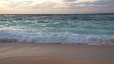 Hawaii 'de gün batımında sakin bir plaj sahnesi. Yumuşak dalgalar, turkuaz su ve kısmen bulutlarla kaplı altın gökyüzü ile kumlu kıyılarda yuvarlanır..