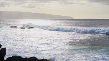 Dalgalar, Hawaii kıyı şeridindeki karanlık volkanik kayalara çarpar. Parçalı bulutlu bir gökyüzü ve uzak dağların altında dalgalar hareket eder..