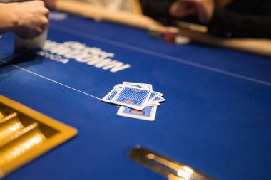 Mavi bir keçeli masa üzerinde poker oynanıyor. Kartlar yüzükoyun, poker fişleri, altın bir çip tepsisi ve kart tutan oyuncular görünür durumda..