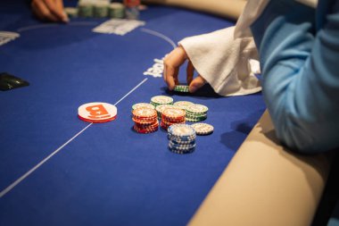 Mavi bir keçeli masa üzerinde poker oynanıyor. Çok renkli çipler, bir dağıtıcı düğmesi ve çip destesine uzanan bir oyuncu eli var..