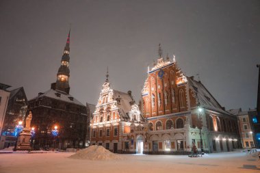 Riga, Letonya 'da karla kaplı. Aydınlatılmış Kara Kafalar Evi, St. Peters Kilisesi kulesi, festival ışıkları ve merkezi bir heykel var..