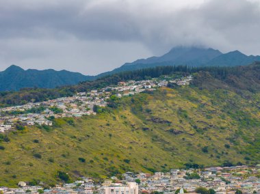 A green hillside with residential homes in Oahu, Hawaii, set against a dramatic mountain range partially covered by low hanging clouds. clipart