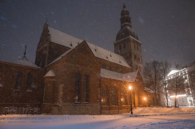 Karlı bir akşamda Letonya 'daki Riga Katedrali' nde. Sıcak sokak lambaları kırmızı tuğlalı Gotik mimariyi aydınlatıyor. Kar yağıyor ve ağaçlar yakınlarda..