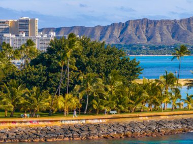 Waikiki kıyı şeridi palmiye ağaçları, turkuaz okyanus, şehir silueti ve arka planda sıcak güneş ışığı altında Diamond Head krateri..