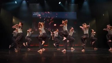 Bir grup çocuk dramatik ışıklandırmayla sahnede dinamik bir dans gösterisi yapıyor. TDB Dans Stüdyosu 'ndan metin gösteren dijital bir ekran.