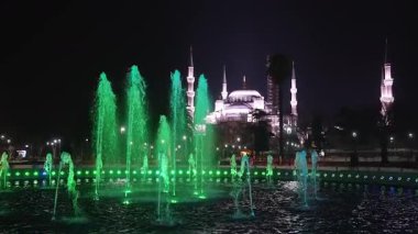 Sultan Ahmed Camii 'nin gece manzarası aydınlık kubbeler ve minareler ön planda renkli bir çeşme ve arka planda yürüyen insanlar..