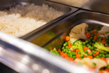 Açık büfe seti, brokoli, karnabahar, bezelye ve havuç da dahil olmak üzere buğulanmış beyaz pirinç ve renkli sebzelerle paslanmaz çelik tepsiler içeriyor..