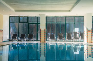 Sakin suyu olan kapalı yüzme havuzu, siyah şezlonglar, yerden tavana orman pencereleri, ve kırmızı ve mavi kiremitli duvarlar..