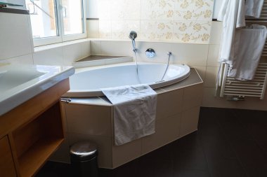 Köşe başında küveti olan modern bir banyo, krem çiçekli fayanslar, ısıtılmış bir havlu askısı, beyaz bornozlar, ve küvetin üstündeki pencereden gelen doğal ışık..