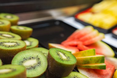 Ön planda dilimlenmiş kivi, arka planda karpuz ve ananas sergilenen canlı bir meyve sergisi, açık büfe ortamında düzenlenmiştir..