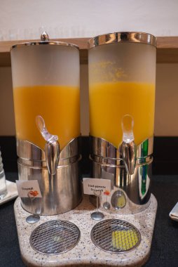 Almanca etiketli benekli bir tezgahta portakal suyuyla dolu iki paslanmaz çelik kap ve yukarıda ahşap bir rafta gözlük var..