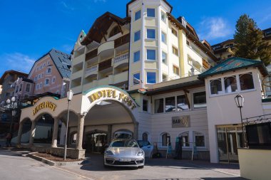 Ischgl 'deki Hotel Post' ta ahşap balkonlu Alp mimarisi, açık sarı cephe ve kemerli girişinin yanına park edilmiş parlak bir araba yer alıyor..