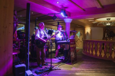 Alpli iki müzisyen gitar ve akordeon eşliğinde sıcacık, ahşap panelli bir alanda, üzerinde Chaluma yazan bir işaret ve ışık ile performans sergiliyorlar..