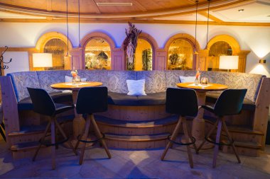 Konforlu oturma alanı yarı yuvarlak ahşap bir kabin, siyah döner sandalyeler, çiçekli yuvarlak masalar ve sıcak aydınlatmalı kemerli ahşap pencereler..