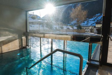 Açık mavi suyu olan kapalı havuz, büyük cam pencereler ve Avusturya 'nın Ischgl kentinde karla kaplı dağlar ve binalar. Güneş ışığı içeri akıyor..