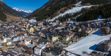 Avusturya 'nın Ischgl kentinde, karlı zirveleri olan bir vadide, sarı bir kilise, kayak pistleri ve geleneksel ve modern binaların karışımı yer alıyor..