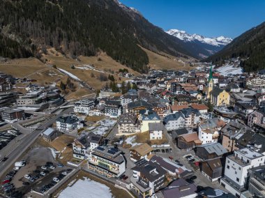 Ischgl, Avusturya 'nın havadan görünüşü, yeşil kulesi olan sarı bir kilise, Alp tarzı binalar, karlı dağlar ve orman yamaçları var..