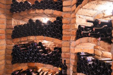 İçinde kırsal tuğla raflar, kemerli tasarımlar ve sıcak ışıklandırma bulunan bir şarap mahzeni. Şişeler düzgünce yerleştirilmiş ve bazı etiketler görülebilir..