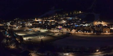 Geceleri Ischgl kayak merkezi, sıcak ışıklar, alp mimarisi, merkezi bir kilise kulesi, karla kaplı yamaçlar ve aydınlık bir park alanı..
