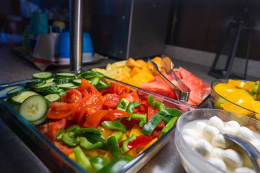 Canlı bir açık büfede, ön planda mozarella topları bulunan salatalık, domates ve karpuz da dahil olmak üzere kesilmiş sebze tepsileri ve meyveler yer alıyor..