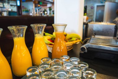 Glass carafes with juices, empty glass cups, fresh bananas, and a stainless steel appliance on a dark countertop in a modern dining area. clipart