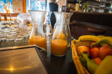 Portakal suyu sürahileri, taze meyve suyu, içme bardakları, minderli koltuklar, bir bar düzeneği, sıcak ışıklandırma ve çiçek duvar kağıdı içeren bir yemek salonu..