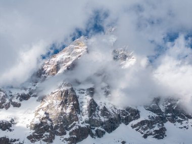 Matterhorn 'lar engebeli, kar kaplı zirveler dramatik bir şekilde yükseliyor, kısmen bulutlar tarafından gizleniyor, güneş ışığı ve gölgeler sivri kaya yüzeylerini vurguluyor..