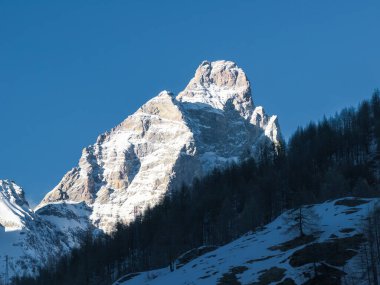 İkonik Matterhorn dağı, kısmen karla kaplı piramidi gibi görünen şekliyle yükselir. Önünde kar parçaları olan bir orman yamacı var..