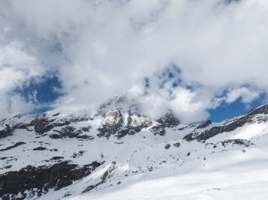 Karla kaplı Matterhorn zirvesi bulutlar tarafından kısmen gizlenmiş kayalık çıkıntılı engebeli arazi ve mavi gökyüzü ve dağınık bulutların karışımı..