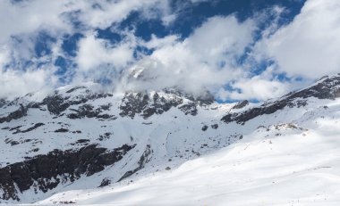 Kar, Matterhorn 'u kısmen bulutlarla kaplamış, raylı ve kayakçılı kayak pistleri ve açık mavi gökyüzünün altında uzak kayak kaldırışları..